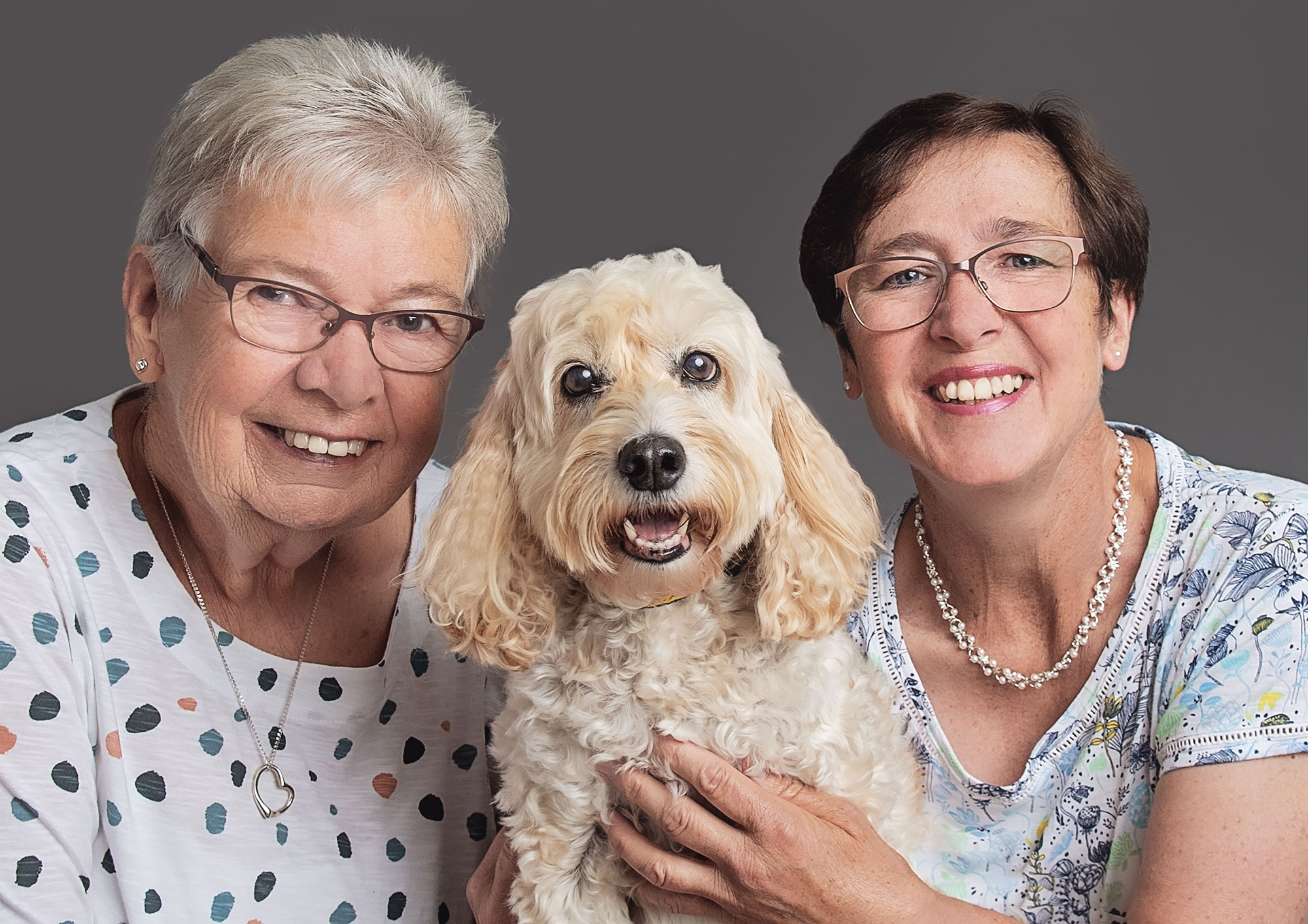 Two smiling elderly women pose with a beige, curly-haired dog, capturing a perfect moment of pet photography. The woman on the left wears glasses and a polka dot shirt, while her friend on the right also sports glasses and a floral shirt. The dog looks forward, appearing happy.